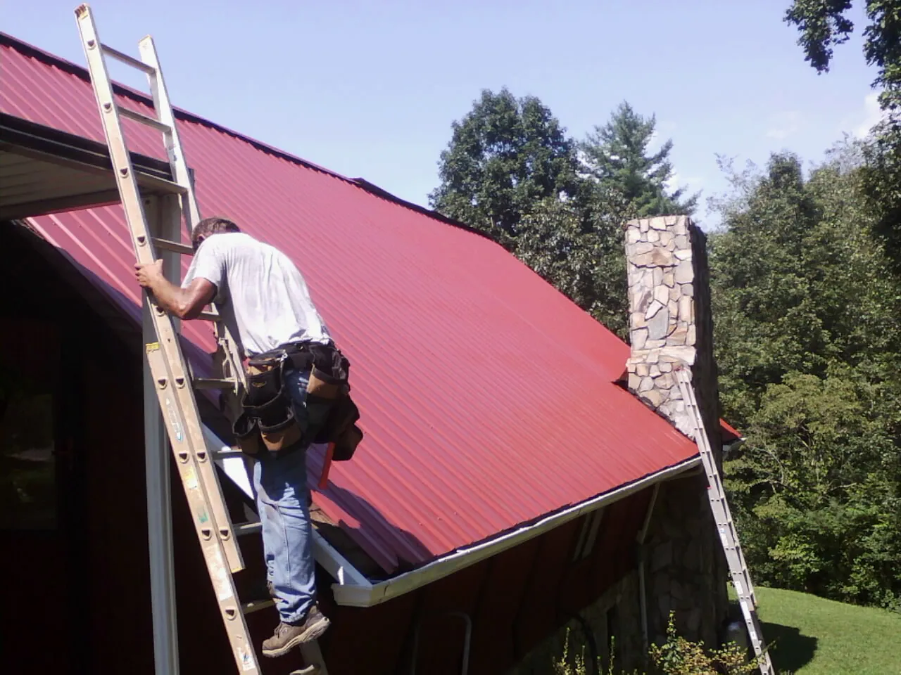 Roofer on a ladder beside a red metal roof and stone chimney — Soffit & Fascia Repair by Roofing Brothers in Karns City